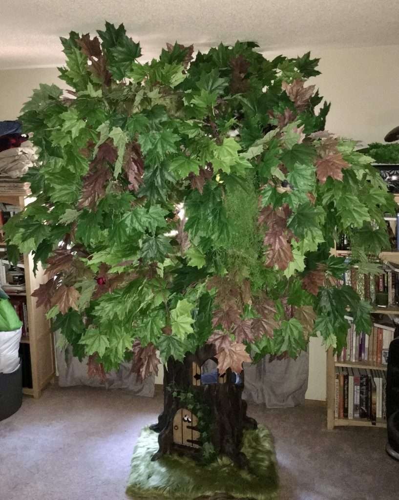 Custom made cat tree with bright green, dark green, brown maple leaves, Spanish moss hanging and faux grass at the base along with hobbit style door on tree trunk and window above. Beige carpet and books in bookcases and light gray curtains in background.