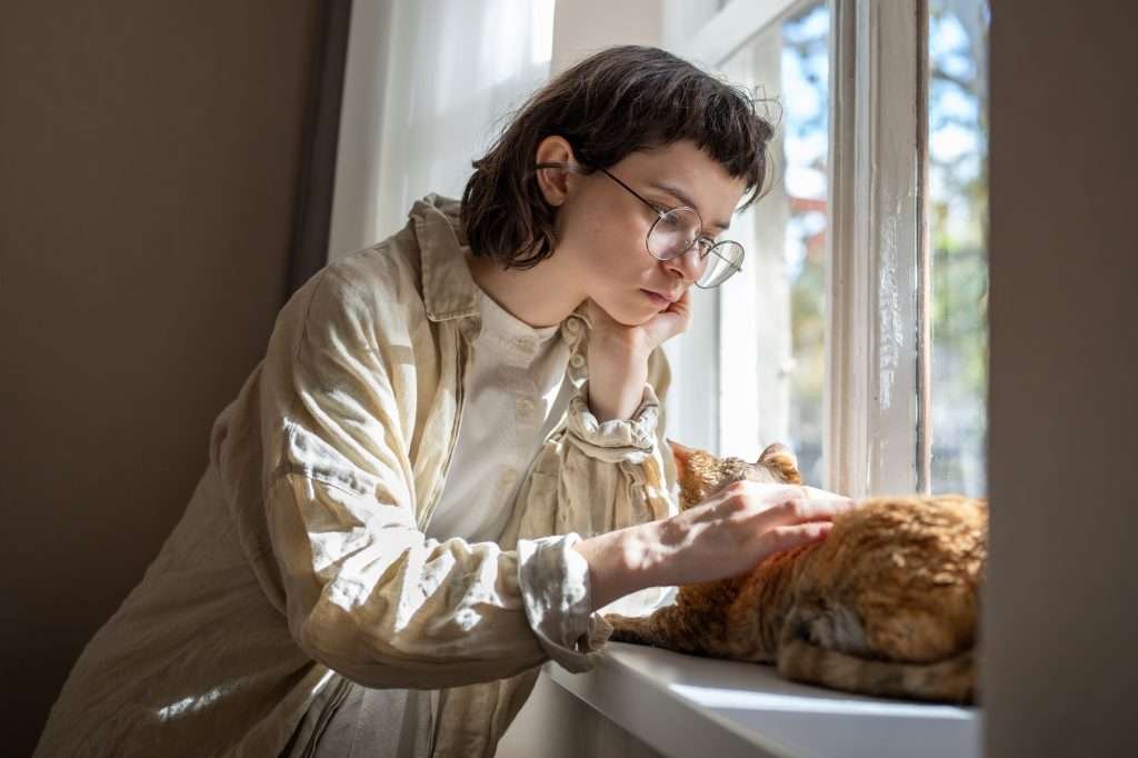 Sad brunette girl wearing glasses and white shirt and a long sleeved beige shirt over it while petting orange tabby cat for emotional support help. Cat is laying on white windowsill in the sun against the window while girl is leaning on hand looking down. Sad brunette girl wearing glasses and white shirt and a long sleeved beige shirt over it while petting orange tabby cat for emotional support help. Cat is laying on white windowsill in the sun against the window while girl is leaning on hand looking down.