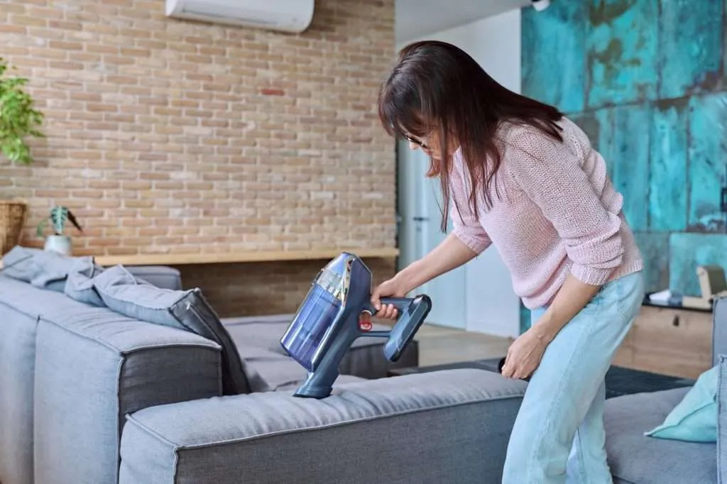 Woman wearing light pink sweater and light blue colored jeans doing house cleaning with handheld blue vacuum cleaner. Brick wall in background with potted plant and teal colored accent wall to the right side. Woman is vacuuming light gray couch.