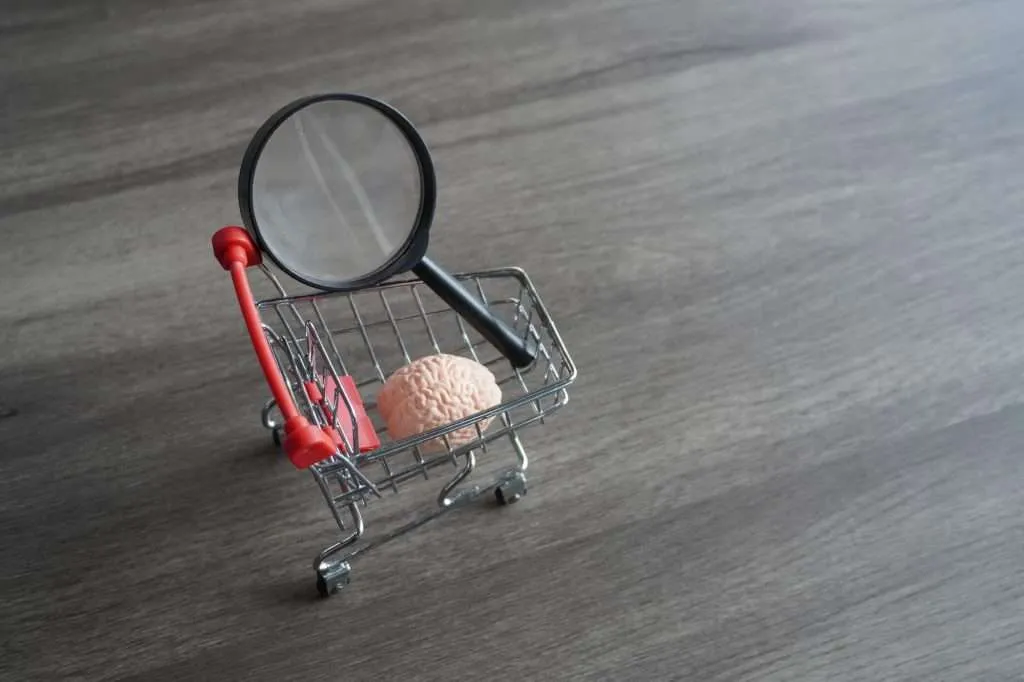 A human brain and magnifying glass inside a mini shopping cart on light wood floor.