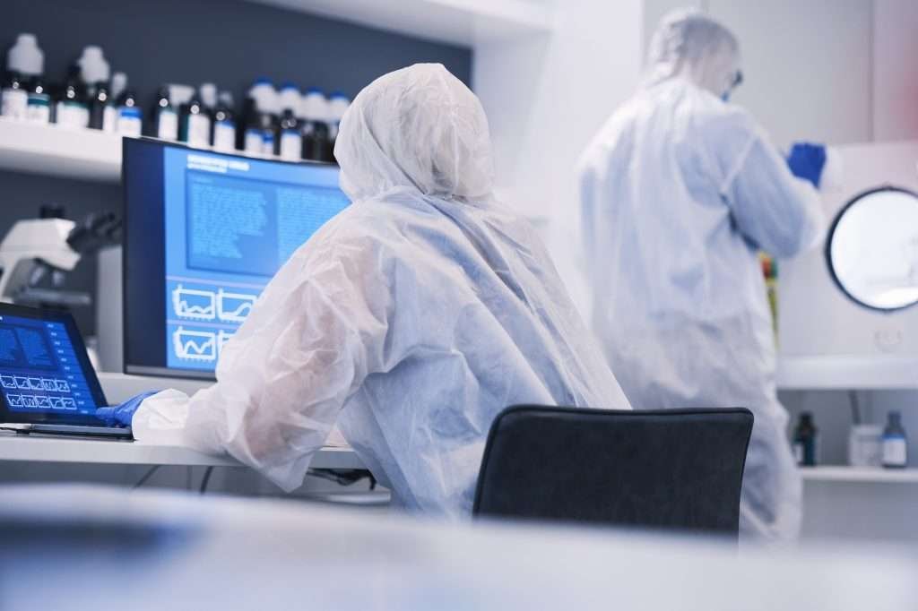Research and science team in an investigation of hazardous virus and in a chemistry laboratory. One person sitting in HAZMAT suit in front of computer and another person standing in HAZMAT suit in background.