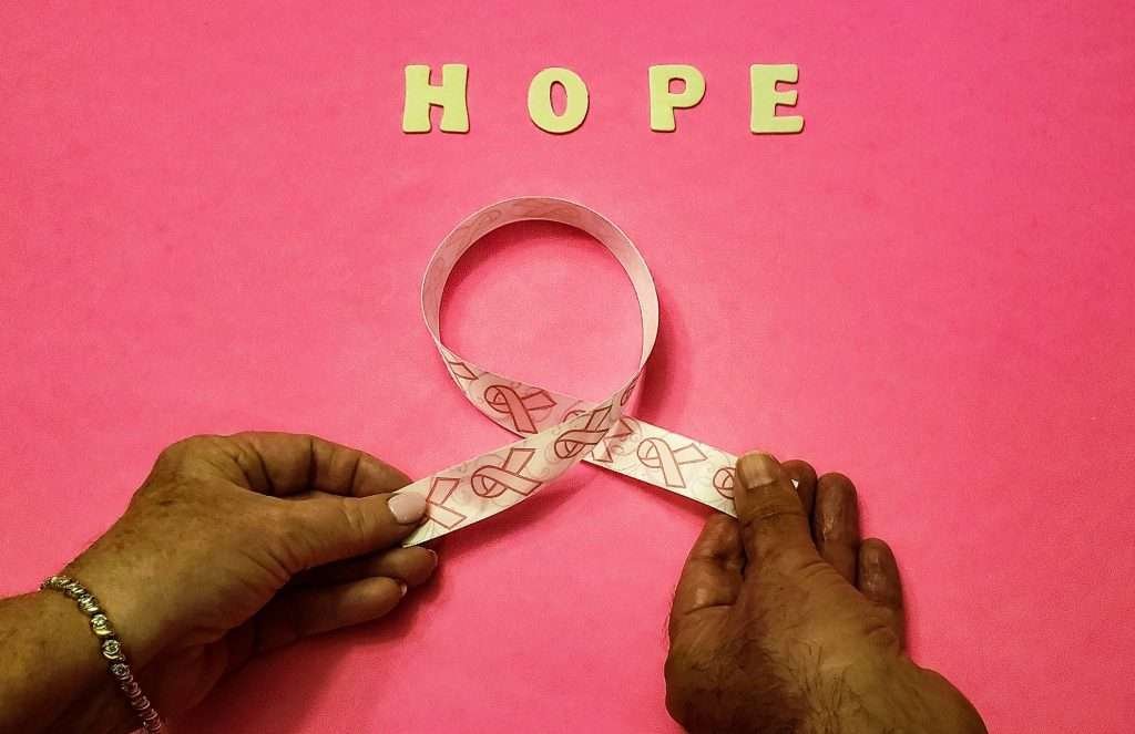 White ribbon with pink ribbons on it being held by two hands and yellow word hope above for the future with bright pink background.