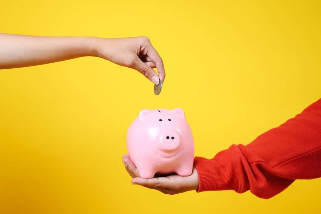 Arm and hand reaching out while putting money into a light pink piggy bank being held by another arm in orange sleeved shirt stretched out holding it.