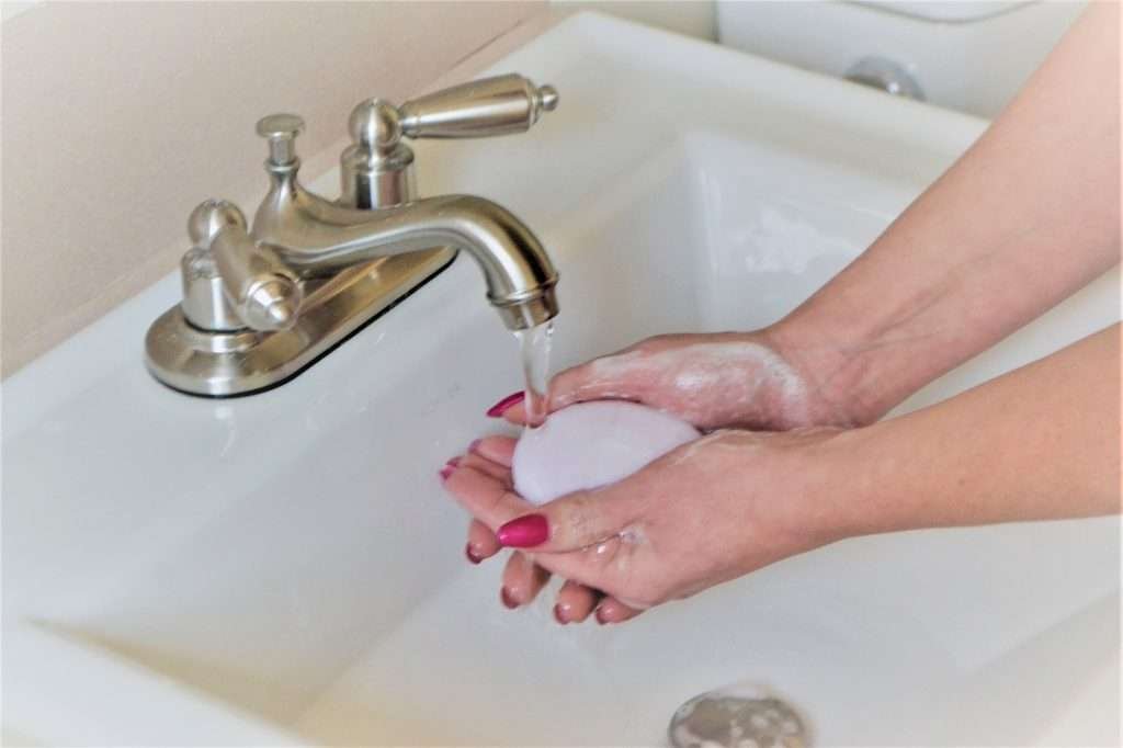 Hand washing with a bar of soap can help prevent illness. Up close - Woman's hands with pink nail polish holding white bar of soap washing under silver faucet in white sink.